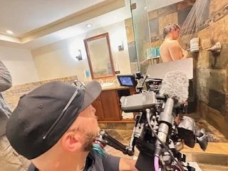 Filmmaker operating camera in a spa or hot tub area, with a person relaxing in the background.