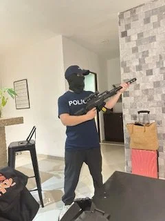 Person in a police uniform holding a rifle, standing indoors in a modern room with a gray stone accent wall and furniture.