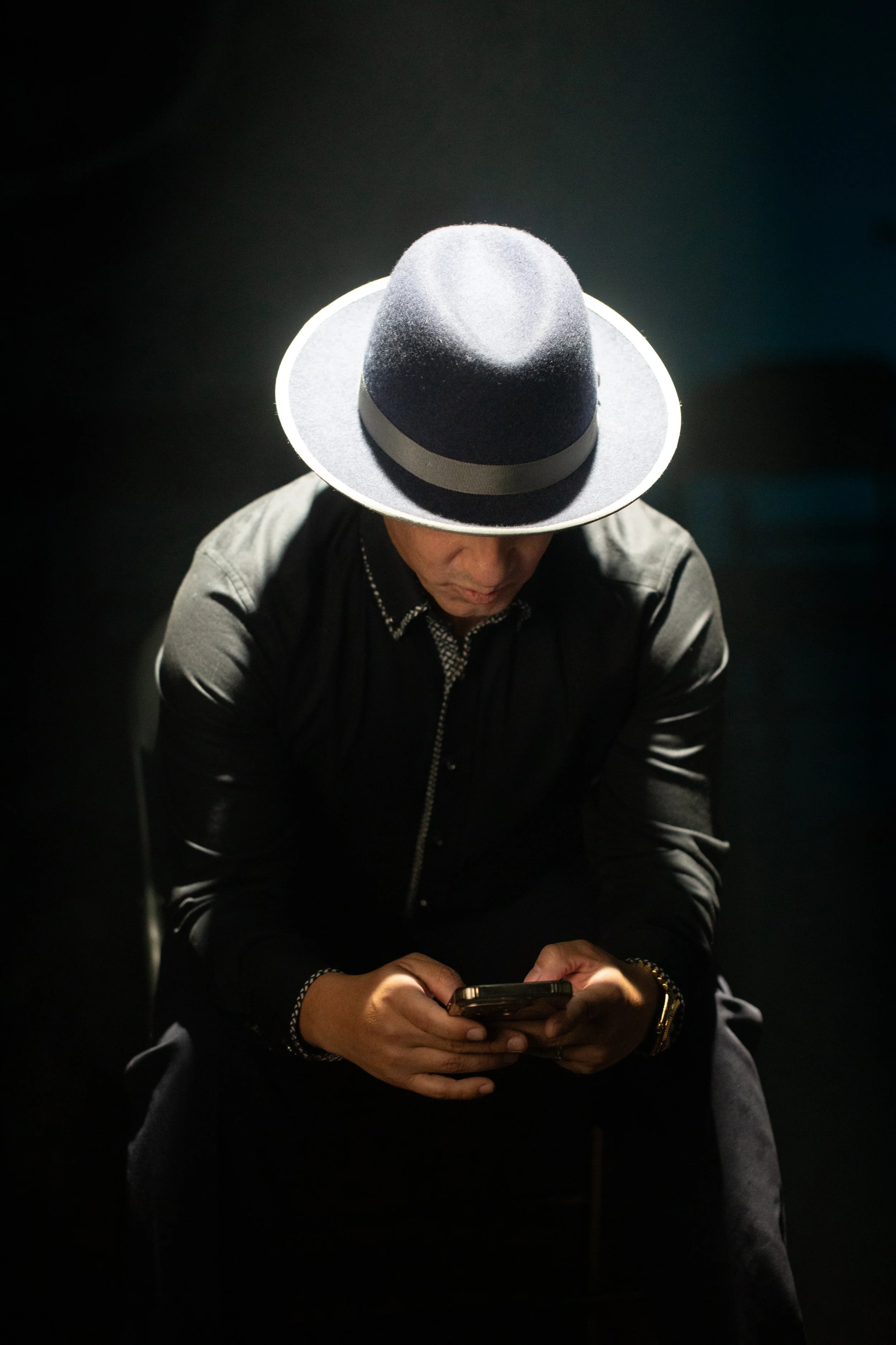 Man wearing a dark shirt and a wide-brimmed hat, sitting in a dark space, looking at his phone.