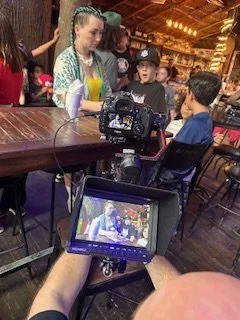 A group of children and teenagers gathered around a table in a restaurant or cafe, crew members are filming them with professional video equipment.