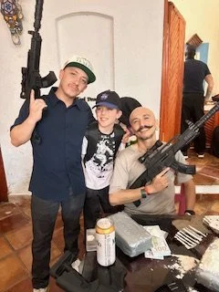 Three men and one young boy posing indoors, holding airsoft guns, with items on a table, in a casual setting.