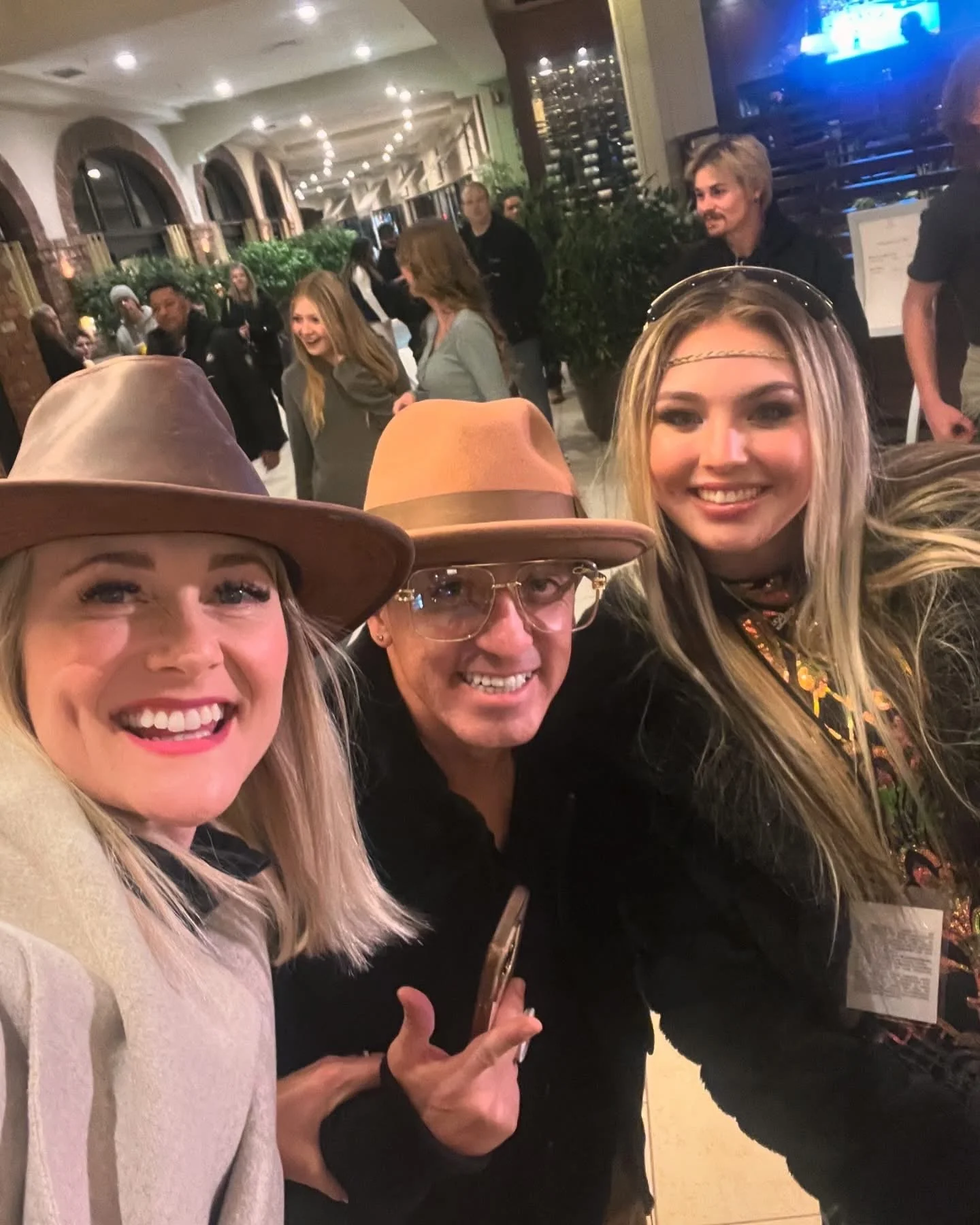 Three friends taking a selfie inside a lively restaurant or bar, smiling and dressed casually with hats, with other patrons in the background.