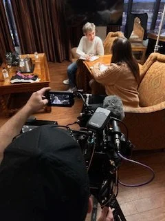 Two women sitting at a table in a cozy indoor setting, being filmed by a person using a professional camera and microphone.