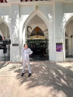 Person in white standing in front of a white arch at an entrance to a building that resembles a palace or mosque, with a sign saying 'Zooroos' above the arch.