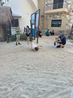 People at the beach, some standing and chatting, one person sitting and taking photos, and a child playing in the sand.