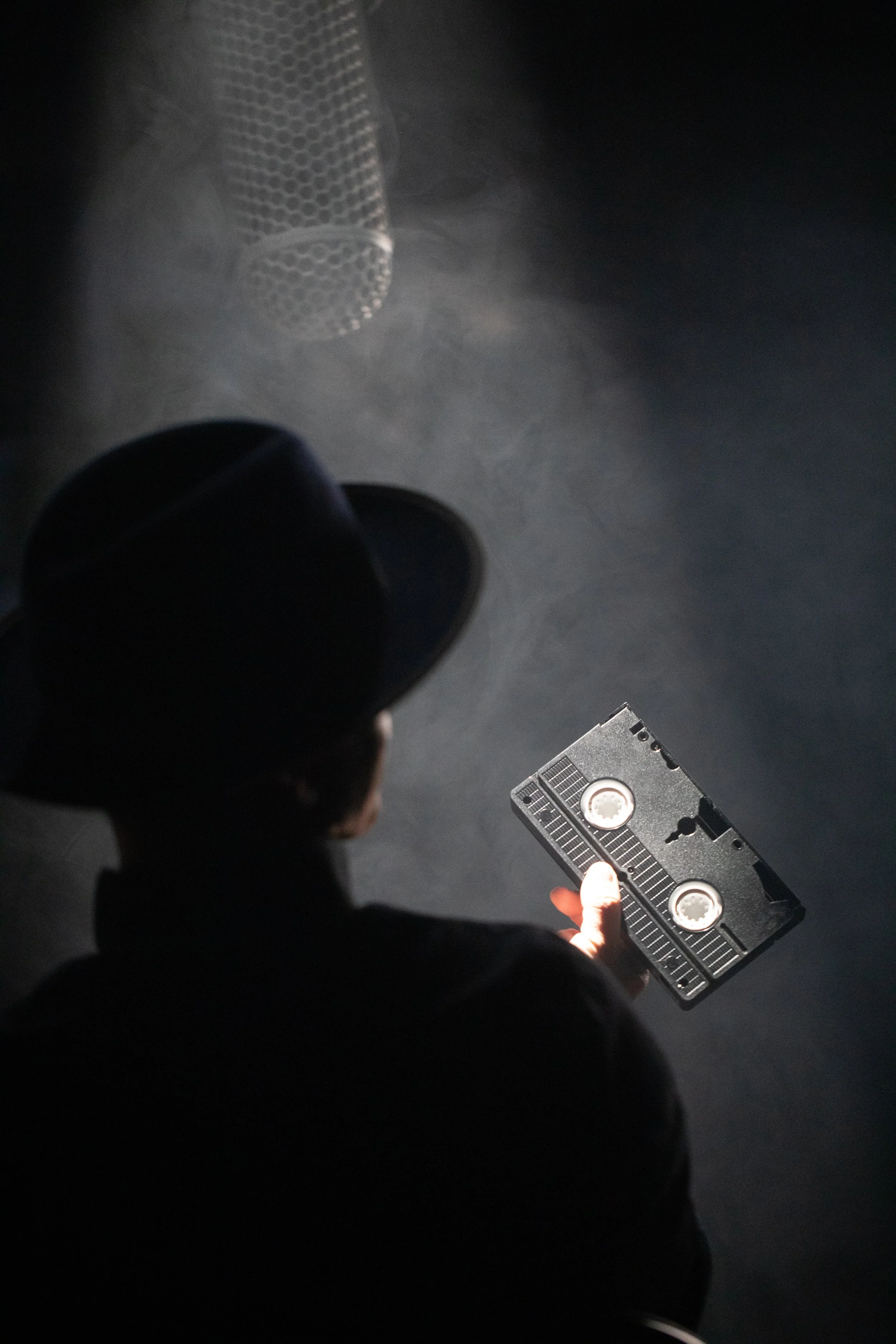 Person wearing a black hat looking at a cassette tape while standing in front of a microphone in a dark studio.