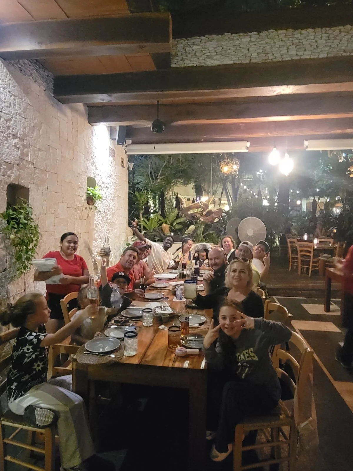 Group of people enjoying dinner at a long wooden table in an outdoor restaurant at night, with plants and trees in the background.