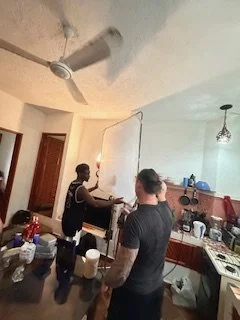 Two men in a kitchen preparing or mixing ingredients, with various items on the countertop and a ceiling fan overhead.