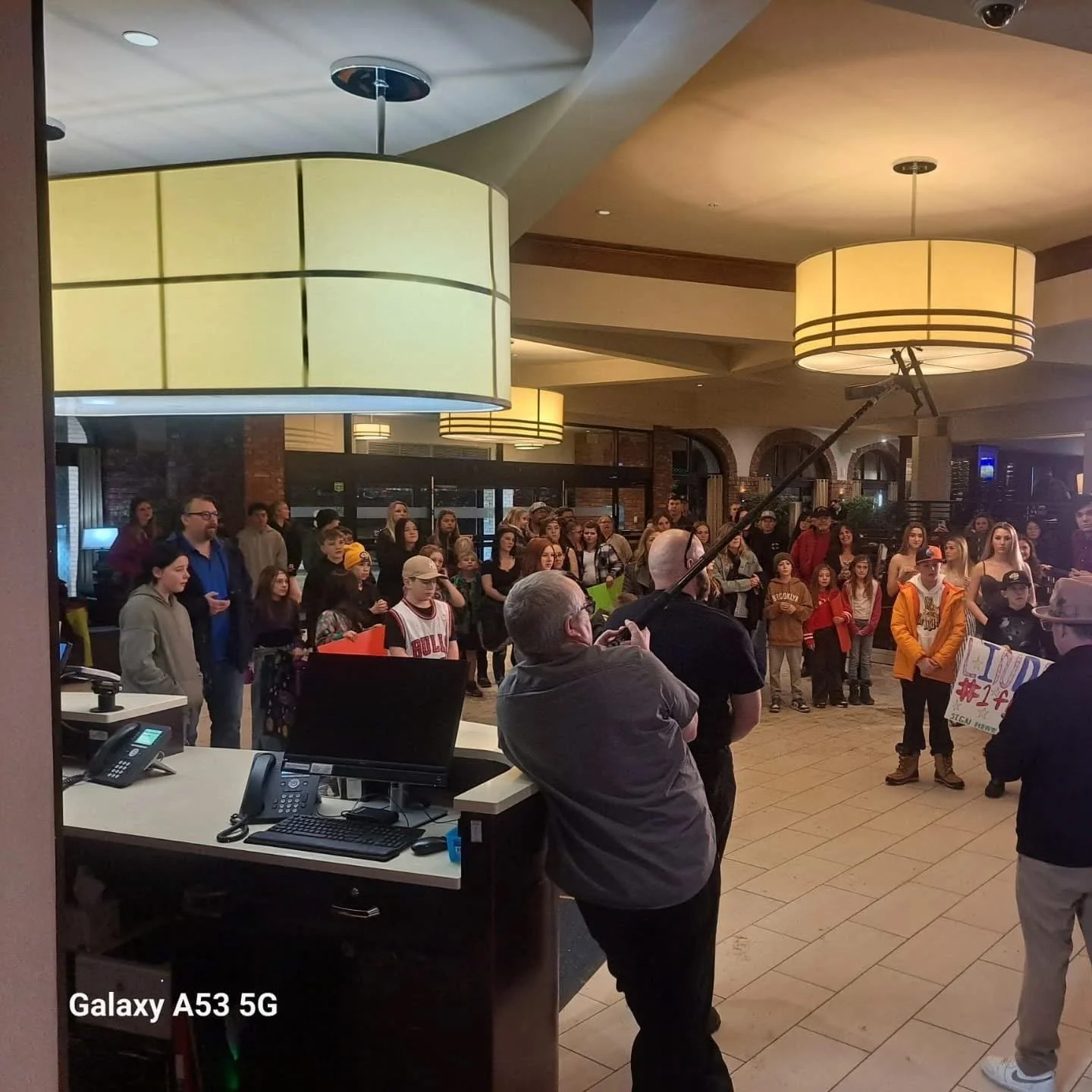 A group of people gathered in a hotel lobby or similar indoor space, with some holding signs or banners, while a person is speaking into a microphone held by a boom pole. Several large ceiling lights are visible, and there are reception desks and com