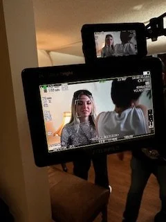 A woman with blonde hair and a headband sitting opposite a person with short dark hair during a filming session, with camera equipment focused on them.