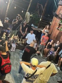 Group of people socializing outdoors, some sitting on chairs, others standing, with a wooden fence and string lights in the background, suggesting a casual gathering or party.