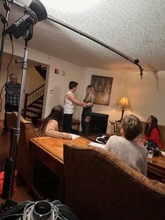 People gathered in a living room with a woman standing and speaking, a person holding a large camera with a boom microphone above them, and others seated around a table.