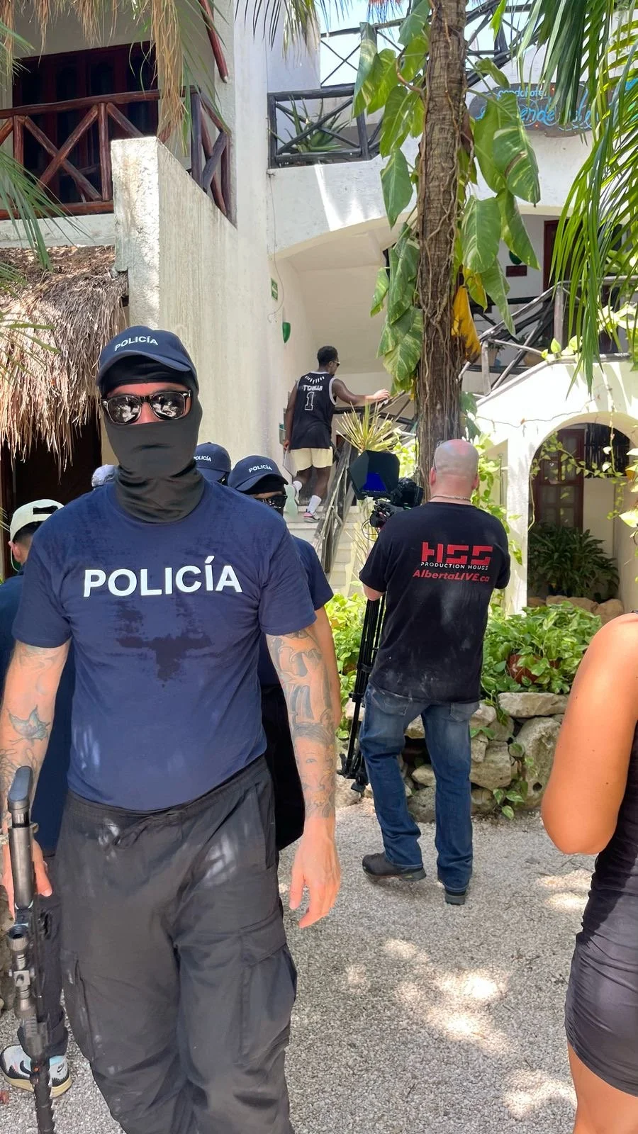 Police officers and a camera crew gathered outside a building with white walls and plants, some people are walking up the stairs, and a man is seen from behind wearing a black sports jersey with the number 1.