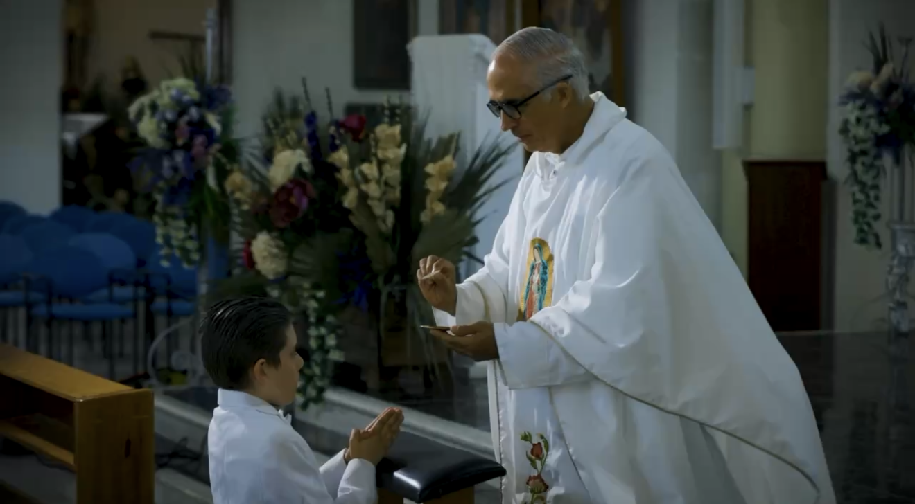 A priest or clergy member in white robes with religious symbols and a young boy in a church, both with their hands clasped in prayer during a religious ceremony.