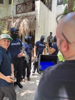 A group of police officers in uniform standing outdoors, some wearing helmets, with a woman smiling and holding a phone in the foreground, near a building and a tree.