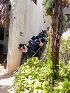 Three police officers aiming their rifles around a corner while taking cover behind a wall outside a building.