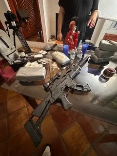 A semi-automatic rifle with a scope and magazine placed on a cluttered table with various objects and a person standing in the background.