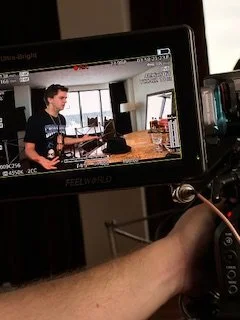 Person sitting at a desk using a laptop, with a window in the background, seen through a camera's viewfinder.