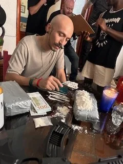 A man is sitting at a table with a cake in front of him, surrounded by friends or family, in a festive setting.