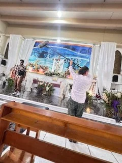 People singing and playing instruments on stage in a church or community hall, with floral arrangements in front and a large screen or backdrop behind them.