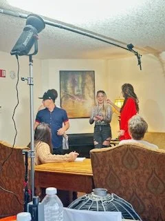 Three women standing and talking in a room with a film crew setup, including a large light and microphone, while two older people sit and watch.