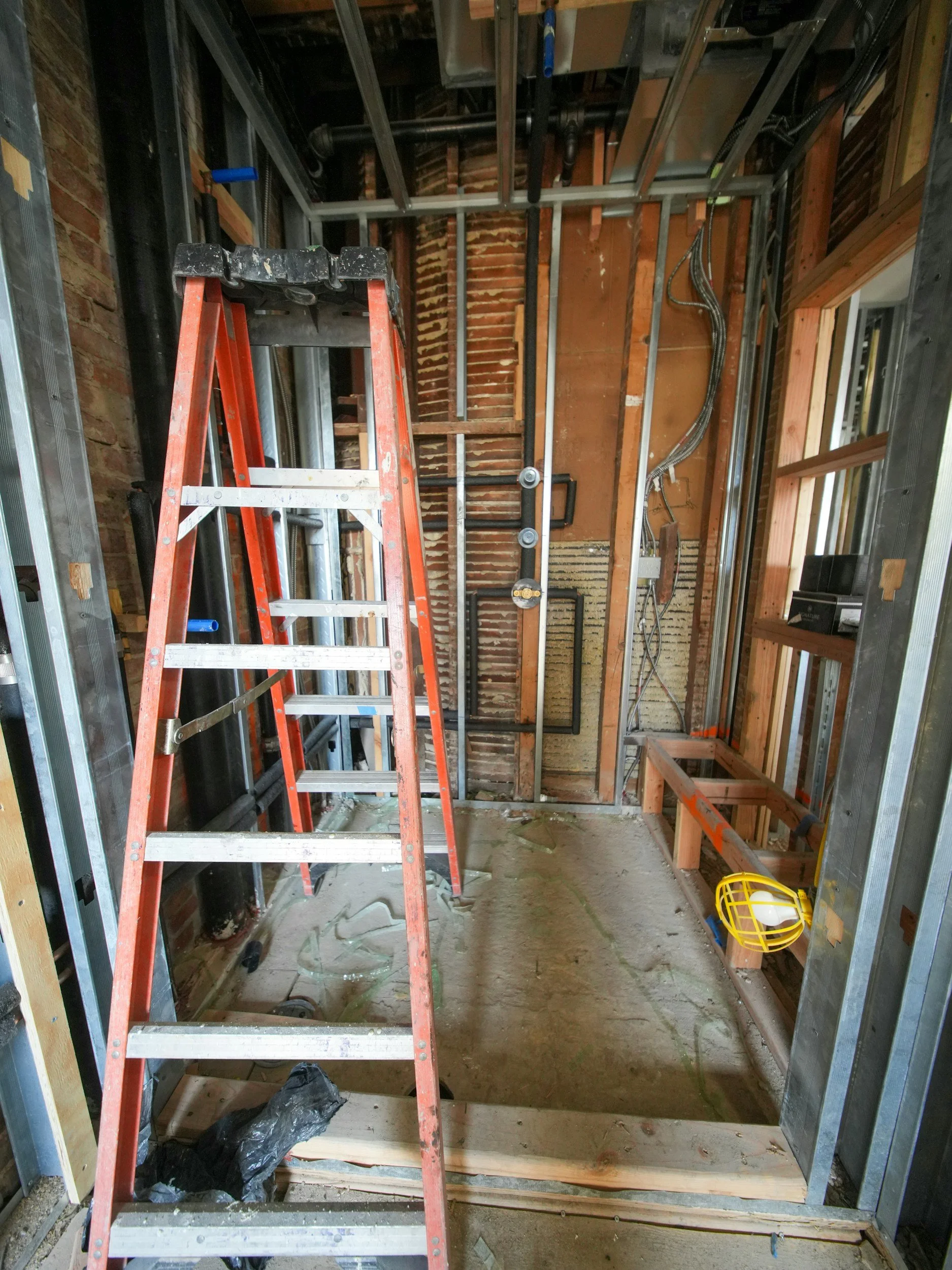 Construction site of a room with exposed brick, wood, and metal framing, a red ladder, and electrical wiring.