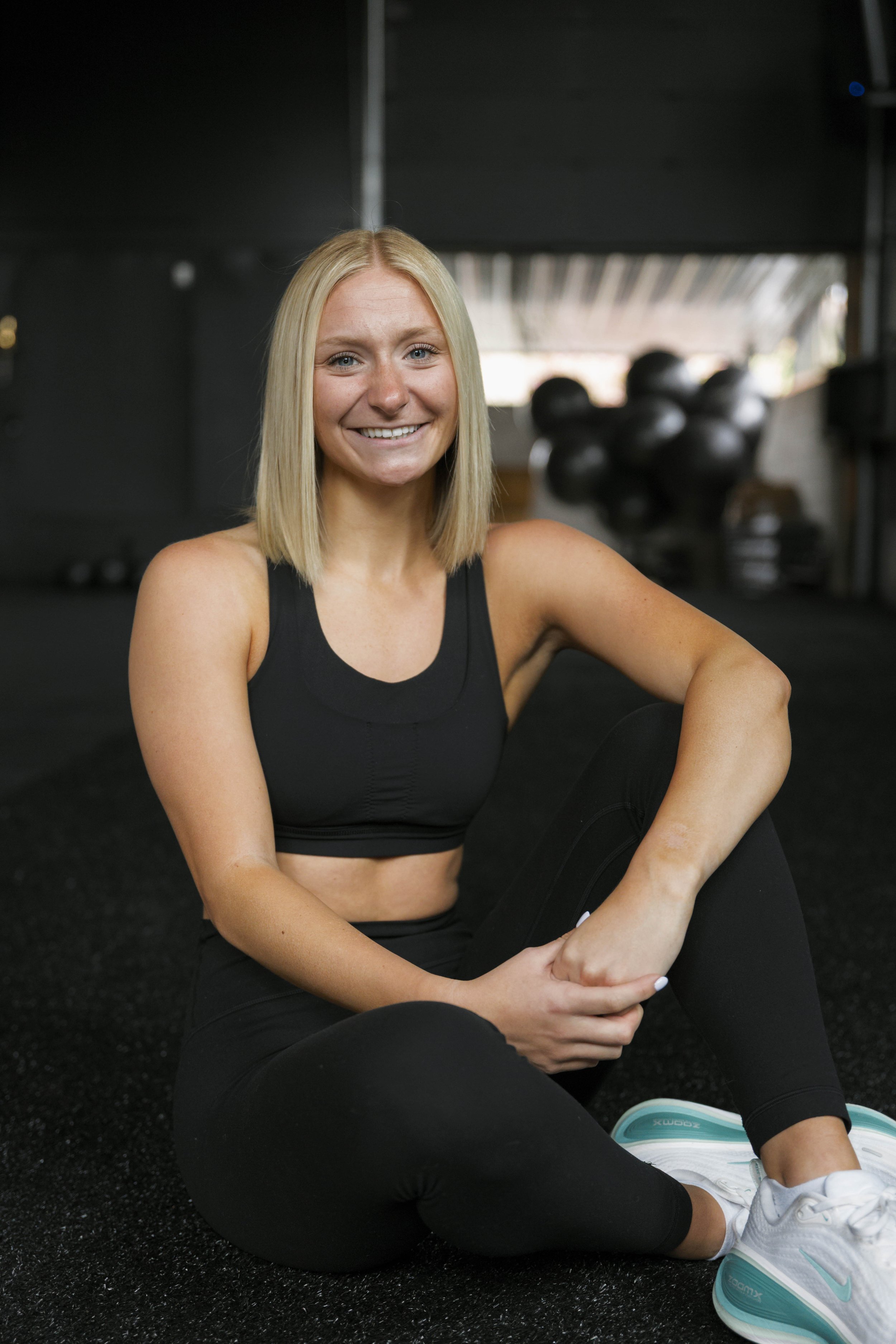 A woman with blonde hair in athletic wear sitting on the gym floor, smiling, with kettlebells in the background.