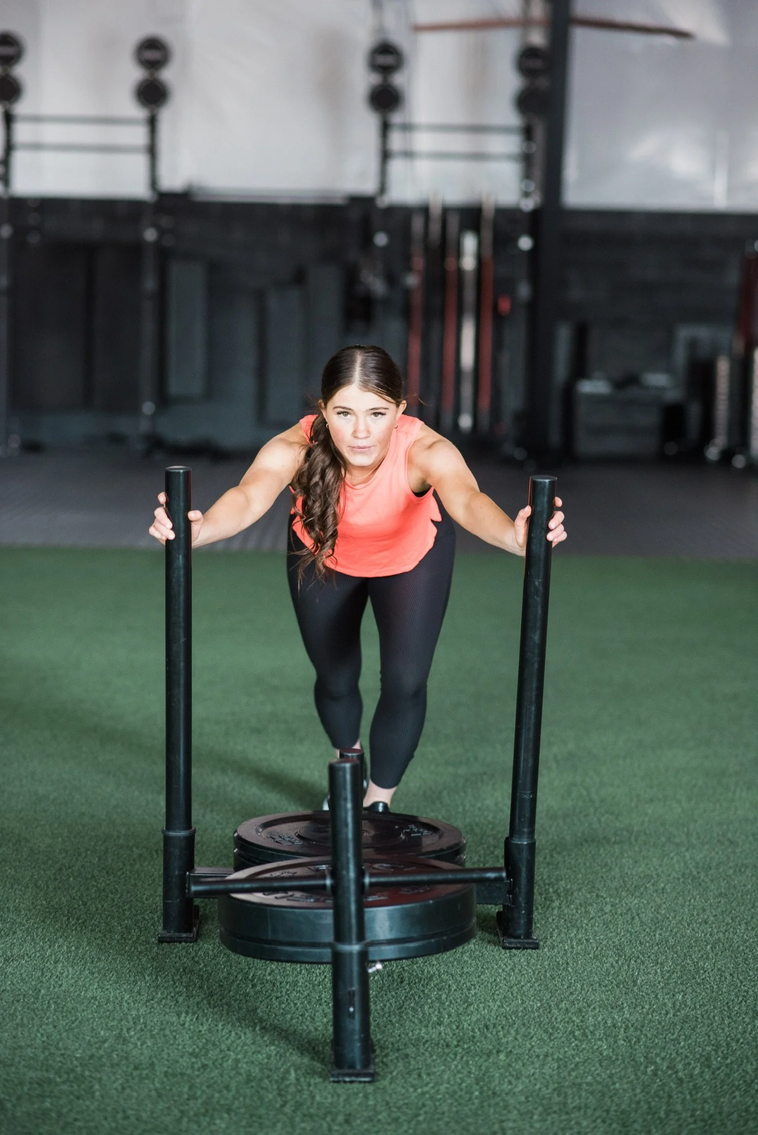 A woman in athletic clothing pushing a weighted sled on a green turf floor inside a gym.