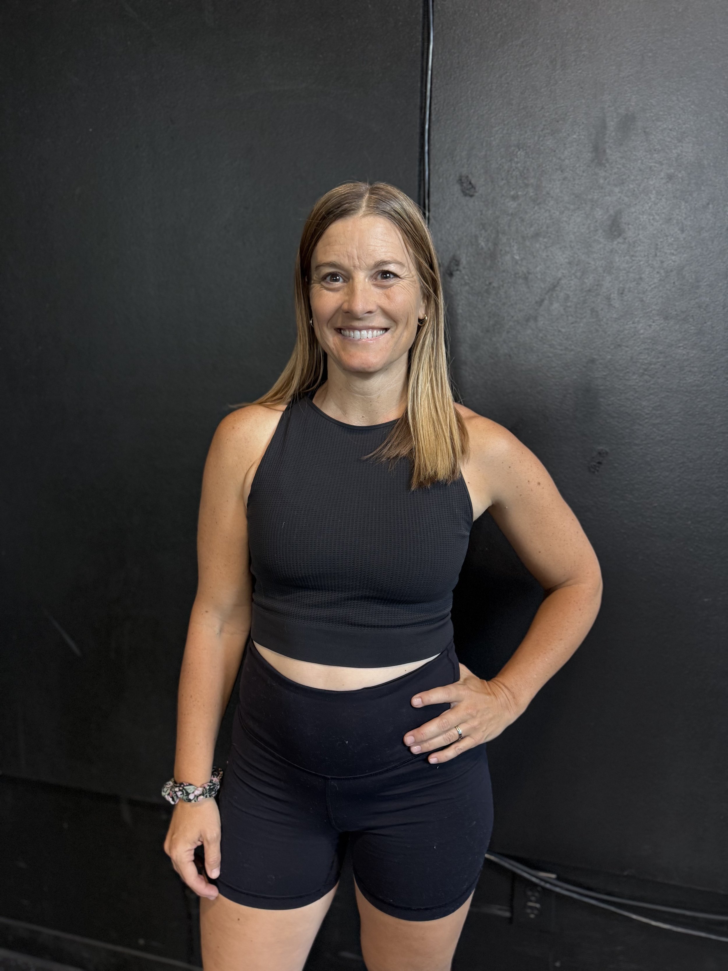 A woman standing in front of a black wall, wearing a black athletic tank top and black shorts, with her right hand on her hip and a smile on her face.