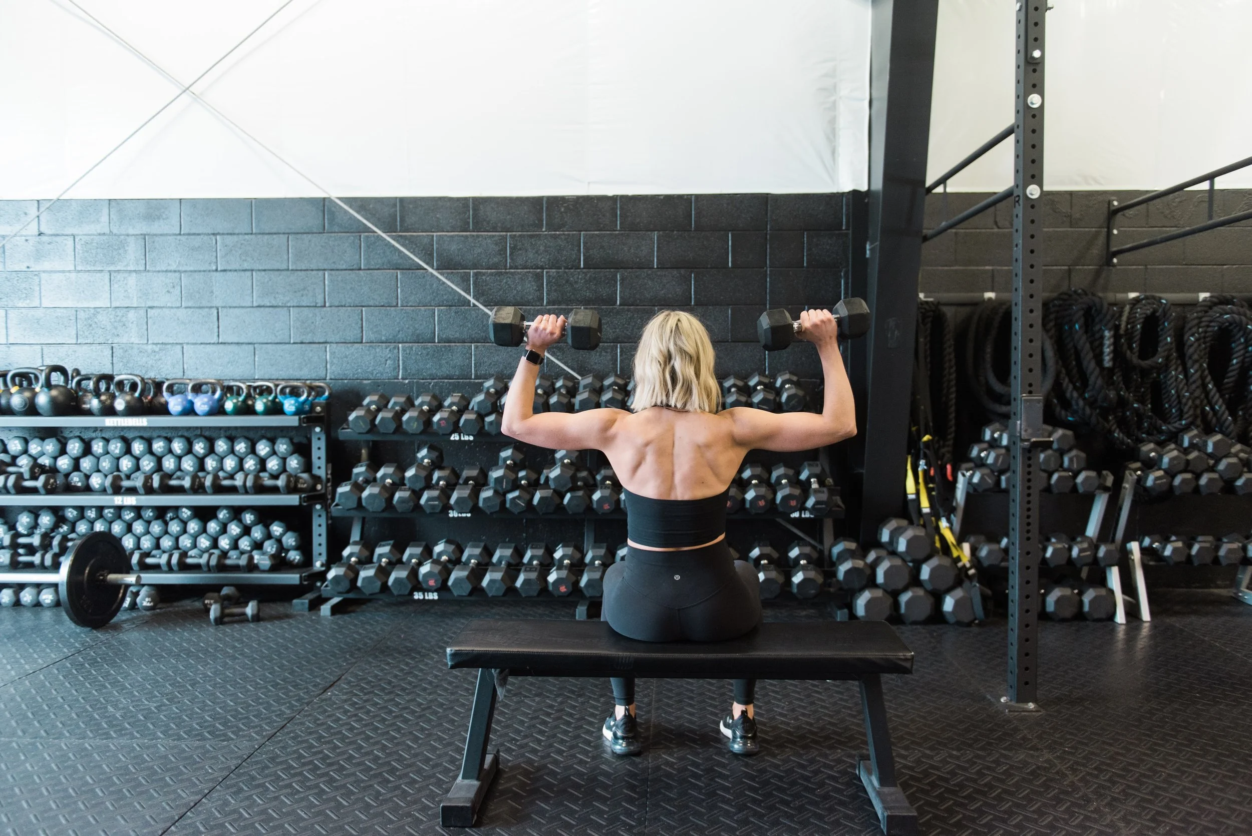 A woman with blonde hair, seen from the back, kneeling on a padded bench, lifting dumbbells at a gym with black brick walls and organized weight racks.