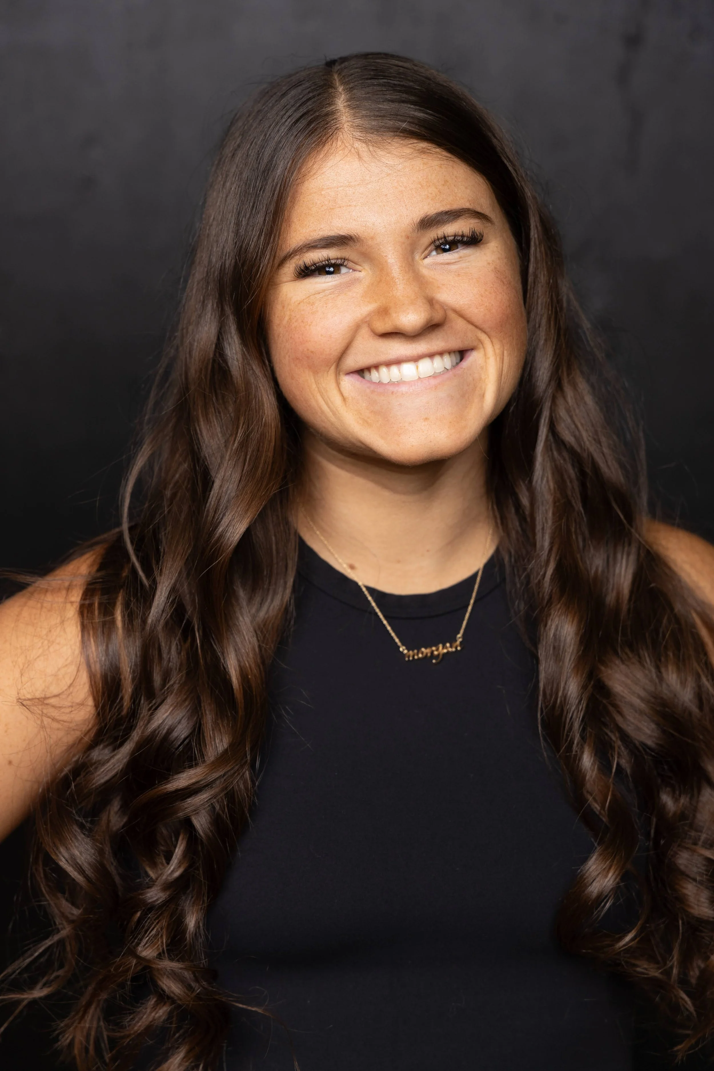 A young woman with long, dark, wavy hair and light skin is smiling on a black background. She is wearing a black sleeveless top and a necklace that says 'morgan'.