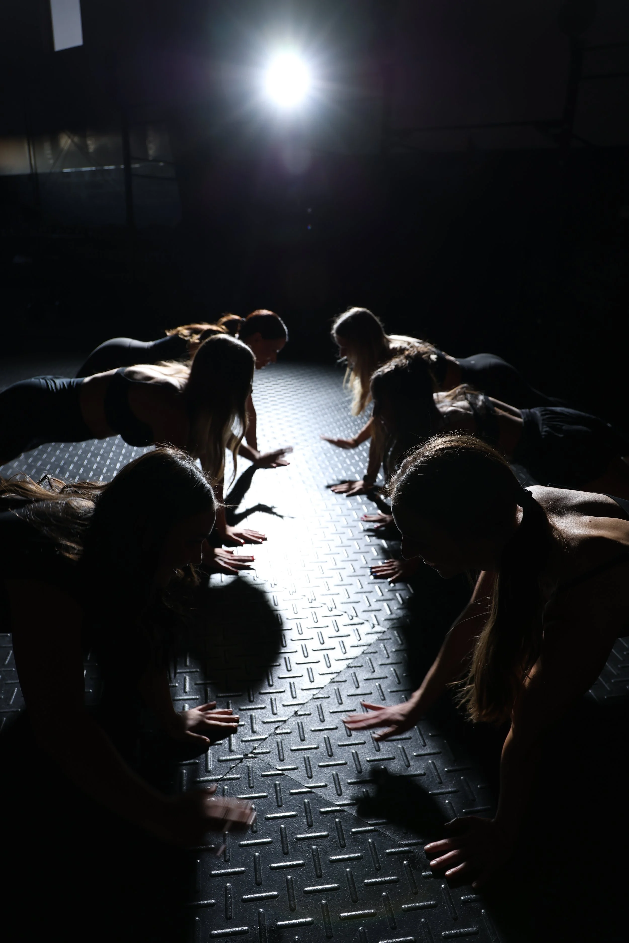 Group of women performing a plank exercise on a textured floor in a dimly lit room with a bright overhead light.