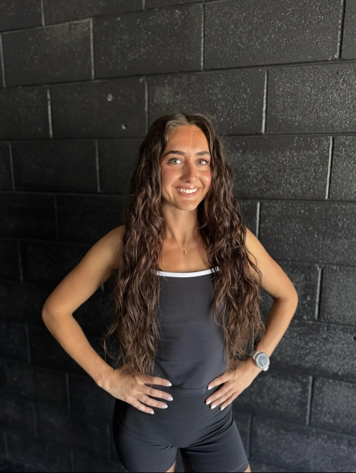 A fit woman with long curly brown hair, blue eyes, and light skin, standing against a black brick wall. She is wearing a black workout tank top with white trim, black workout shorts, a wristwatch, and a small necklace. She is smiling and has her hands on her hips.