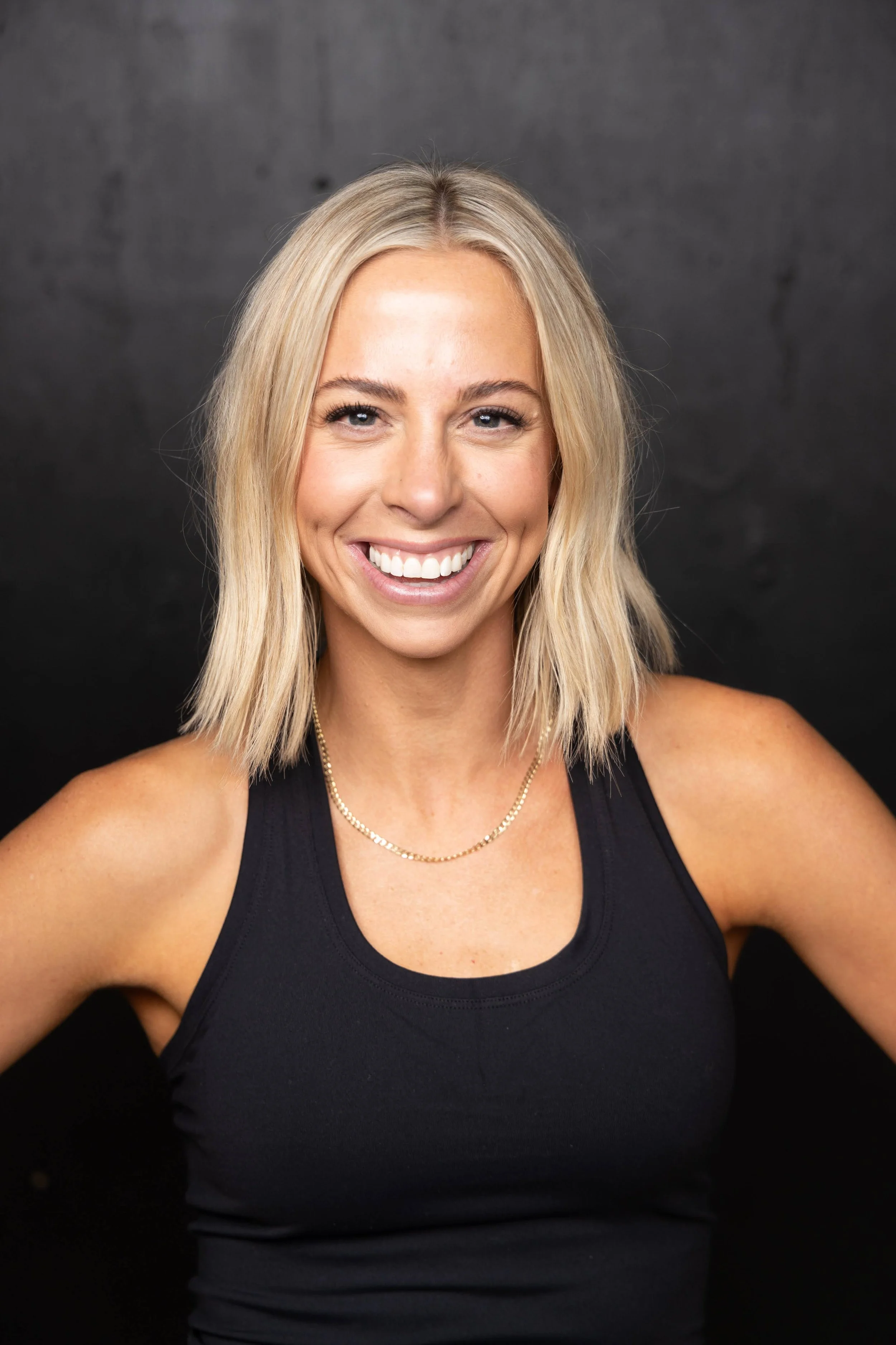A woman with blonde hair and a bright smile, wearing a black tank top and a gold necklace, posing against a black background.
