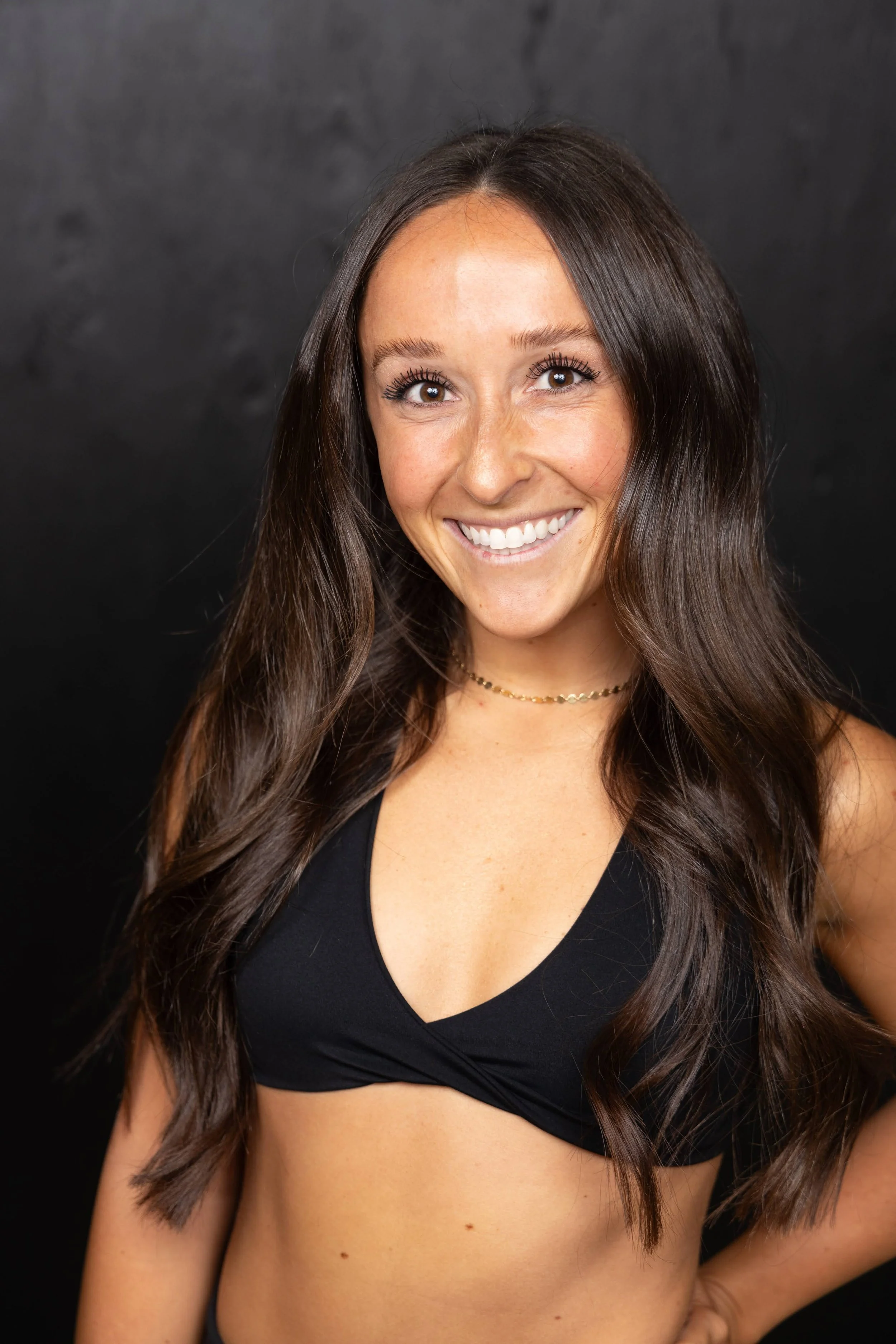 A woman with long wavy brown hair, smiling, wearing a black top and a gold necklace, against a black background.