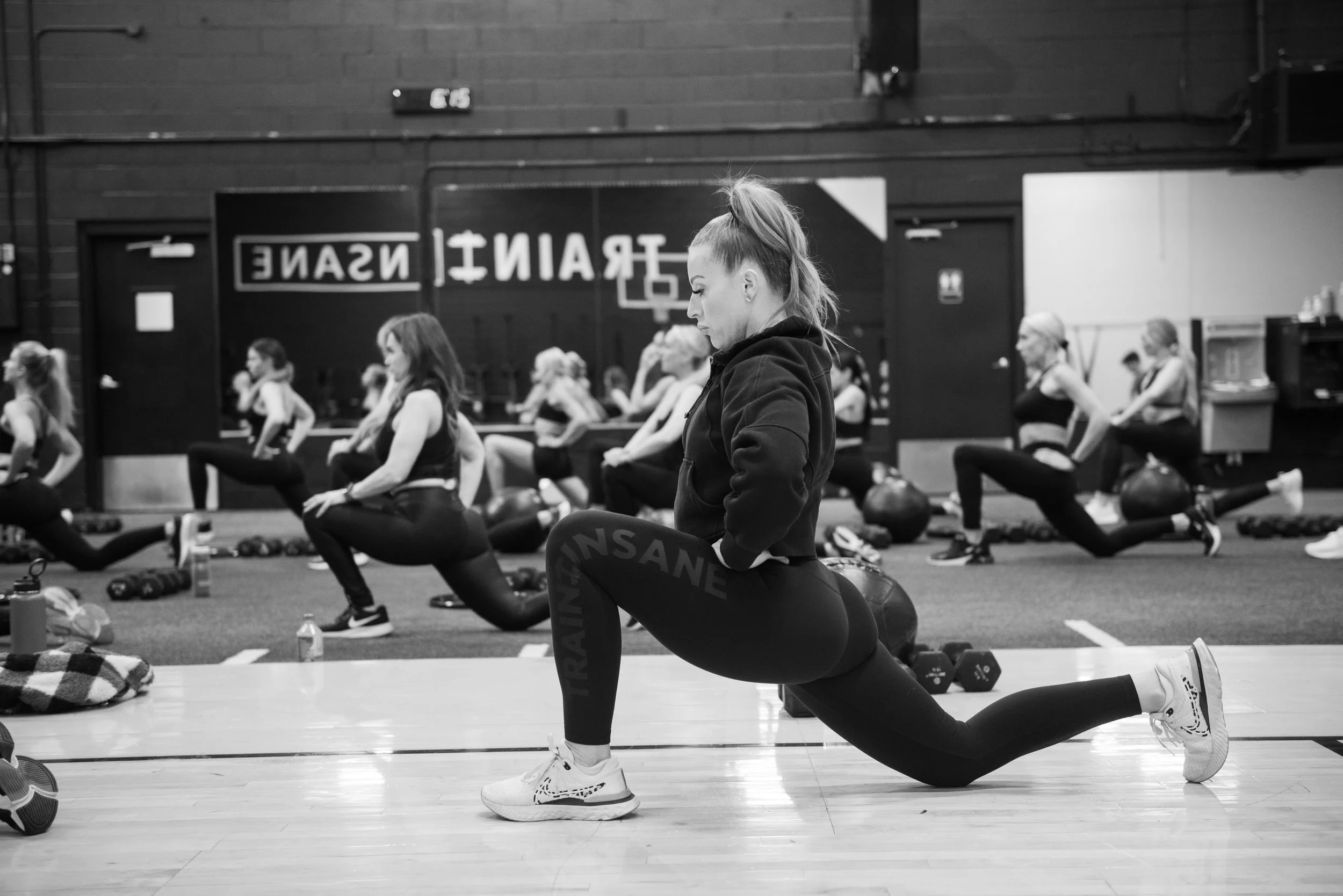 A fitness instructor demonstrating a lunge exercise to a group of women in a gym class, all wearing workout clothes and sneakers, with gym equipment and a large sign in the background.
