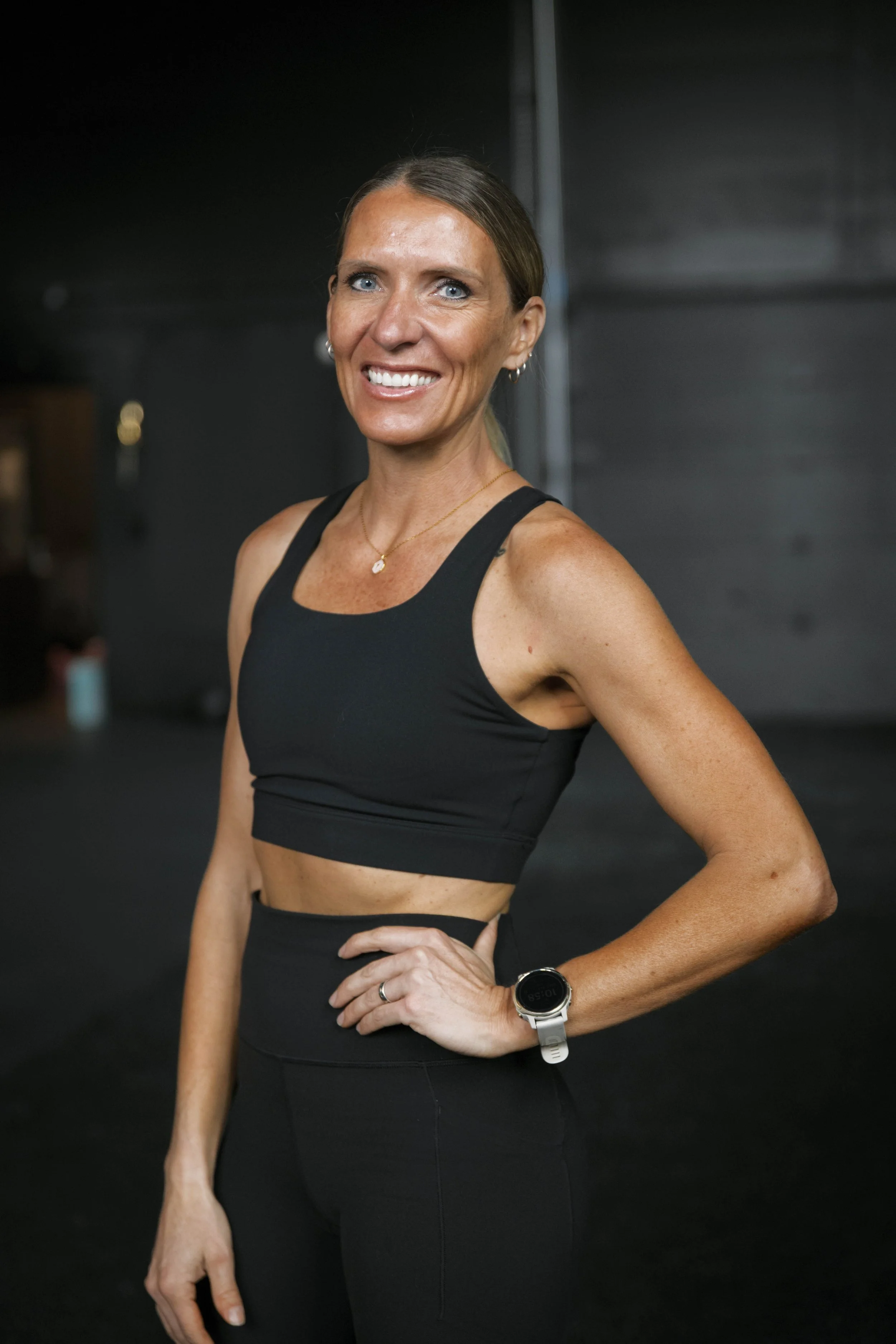 A woman smiling with blue eyes, in a black athletic outfit, wearing a watch and jewelry, standing in a gym with a dark background.