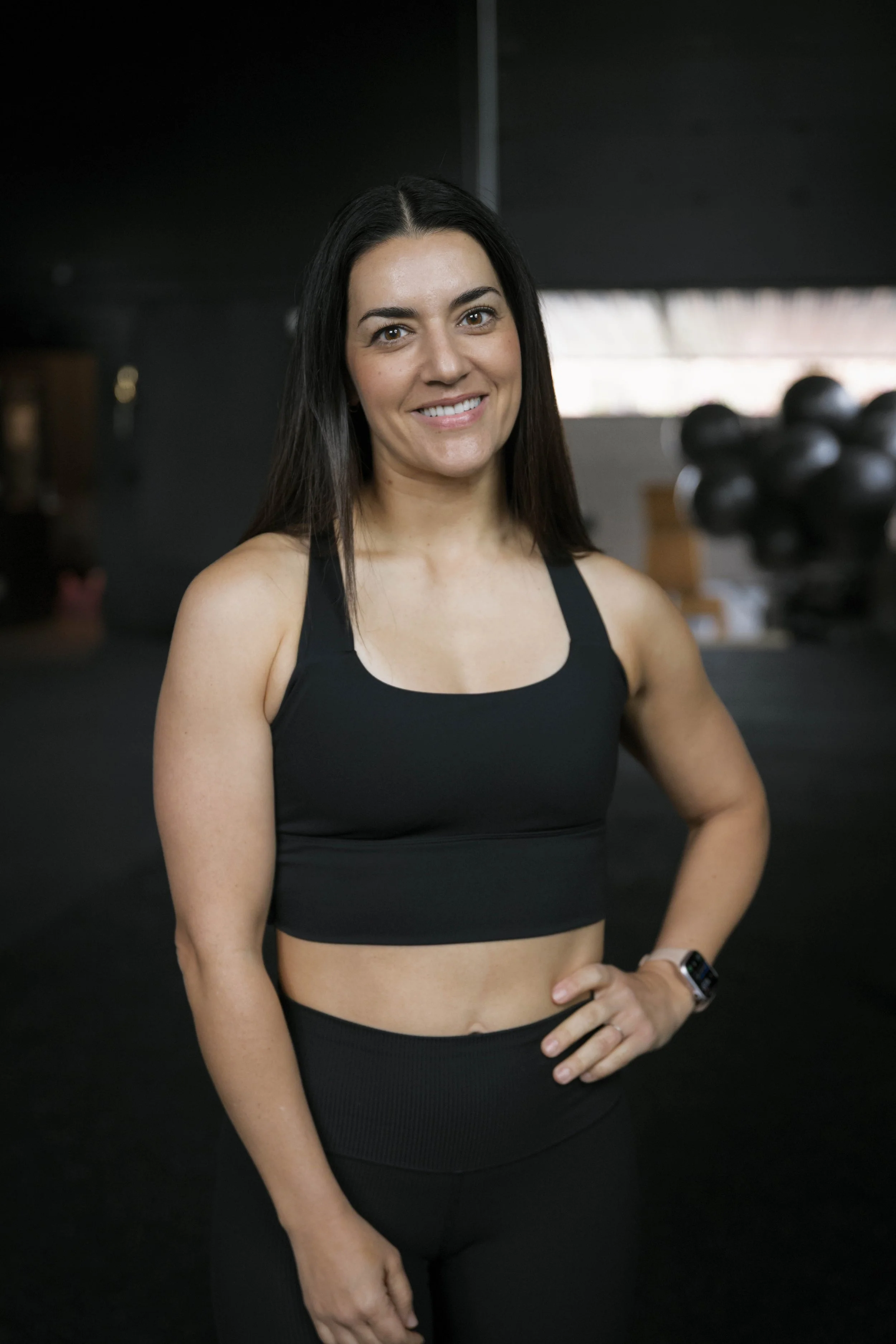 A woman with long dark hair, smiling, wearing a black workout top and black workout pants in a gym. Her left hand rests on her waist, and she is wearing a smartwatch on her left wrist. There are black balloons in the background.