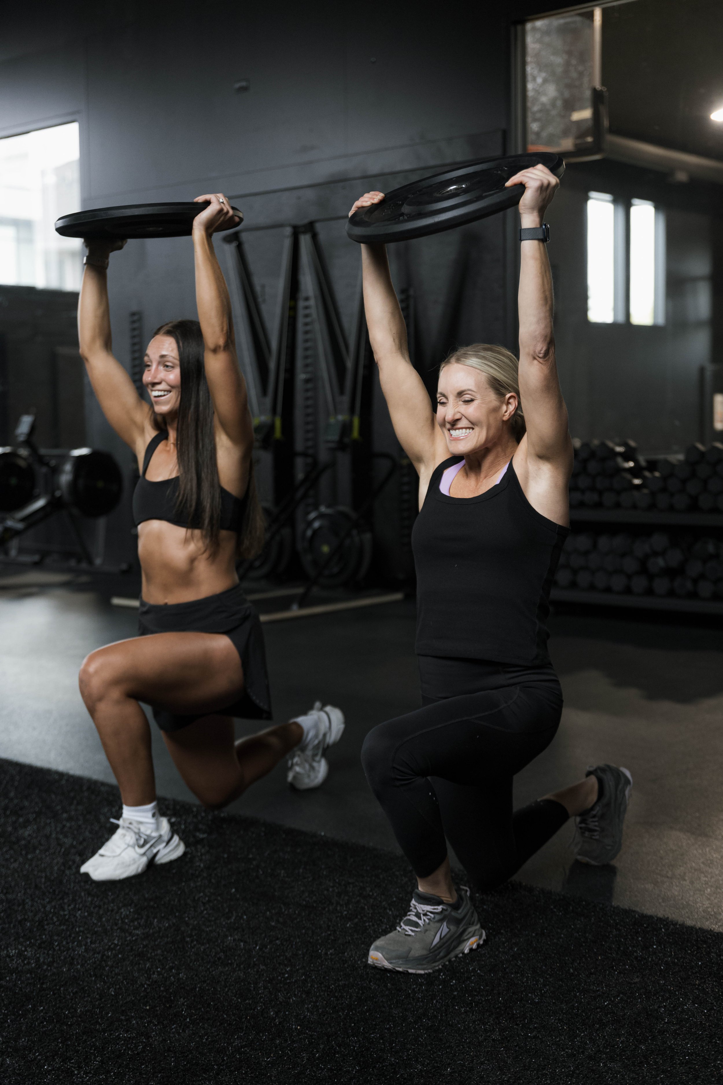 Two women with athletic builds exercising in a gym, lifting weights labeled with weight plates, and kneeling on one knee on a black workout mat. Both women are smiling and wearing workout clothes and sneakers.