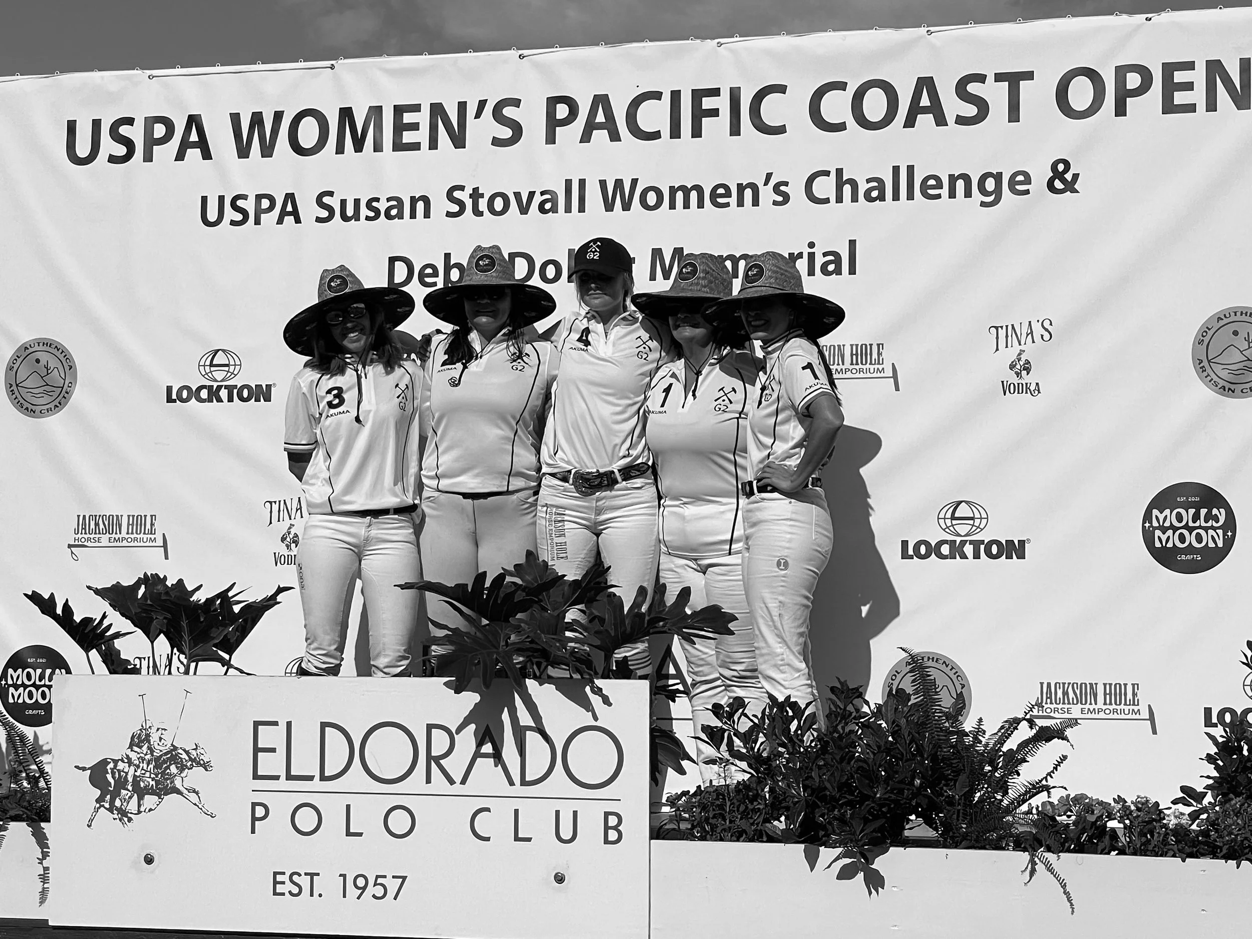 Group of five women posing on a podium at the USPA Women's Pacific Coast Open, wearing matching polo shirts and wide-brimmed hats, with a banner in the background, and plants in front.