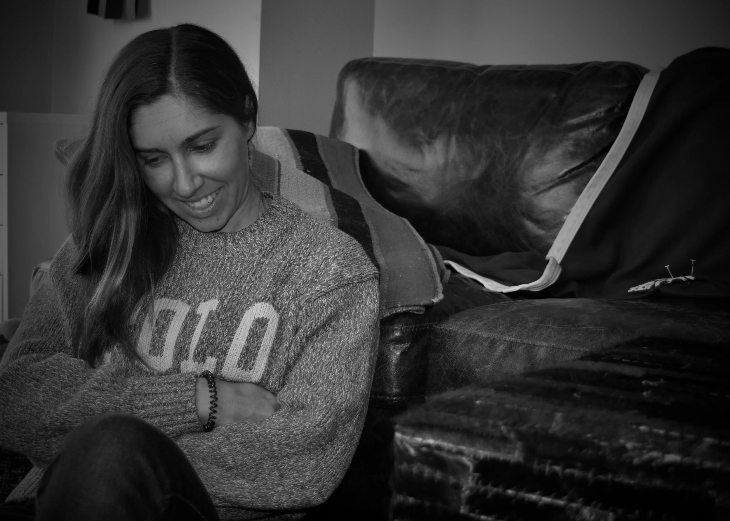 A woman smiling and leaning on her crossed arms, sitting in a room, with a couch nearby, captured in black and white.