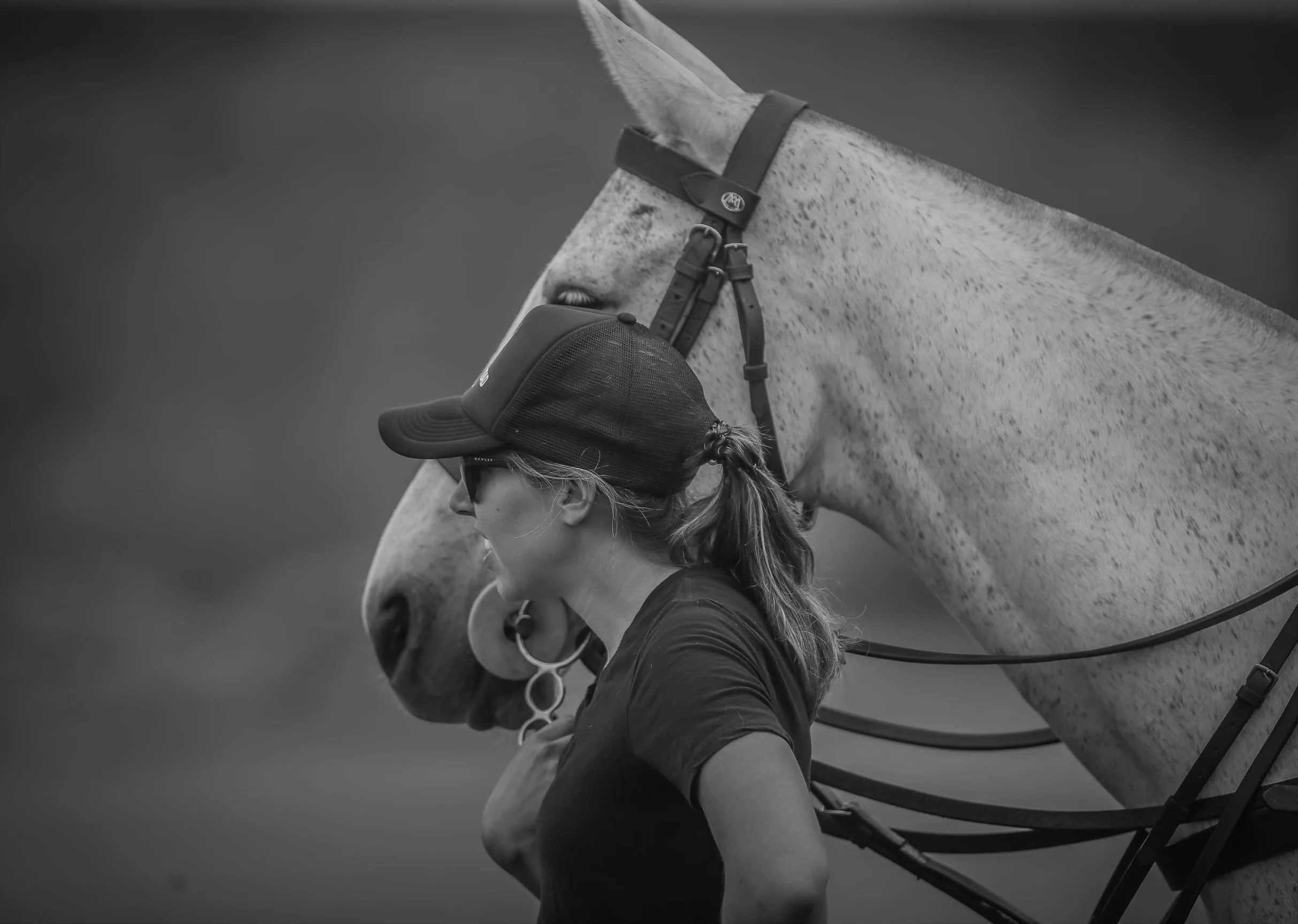 A woman in a baseball cap and sunglasses walks alongside a white polo pony with a speckled coat, which is tacked up ready to play a match