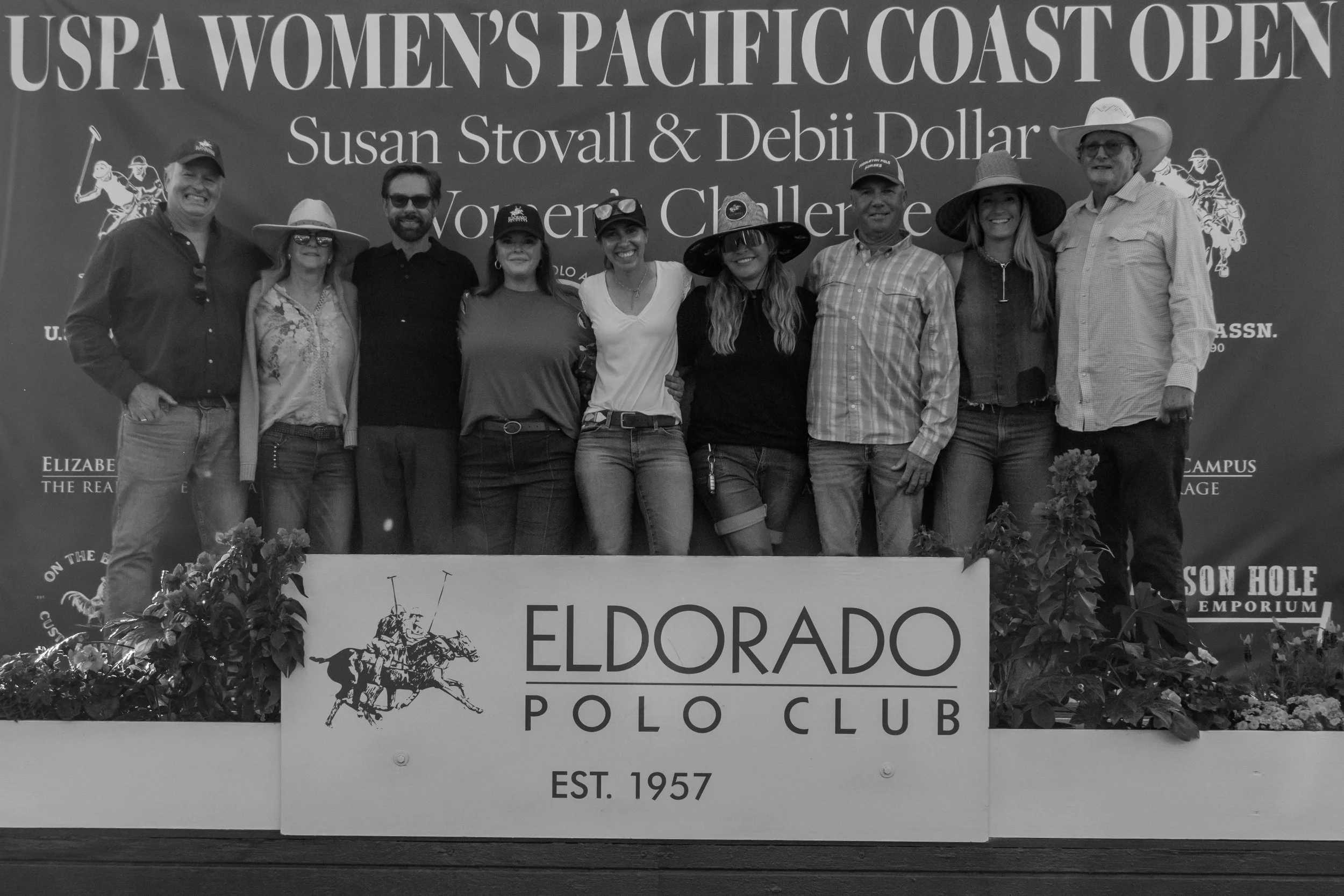 A group of nine people standing together on a podium at the USPA Women's Pacific Coast Open, with a banner behind them. The group is dressed casually, some wearing hats, and they are smiling.