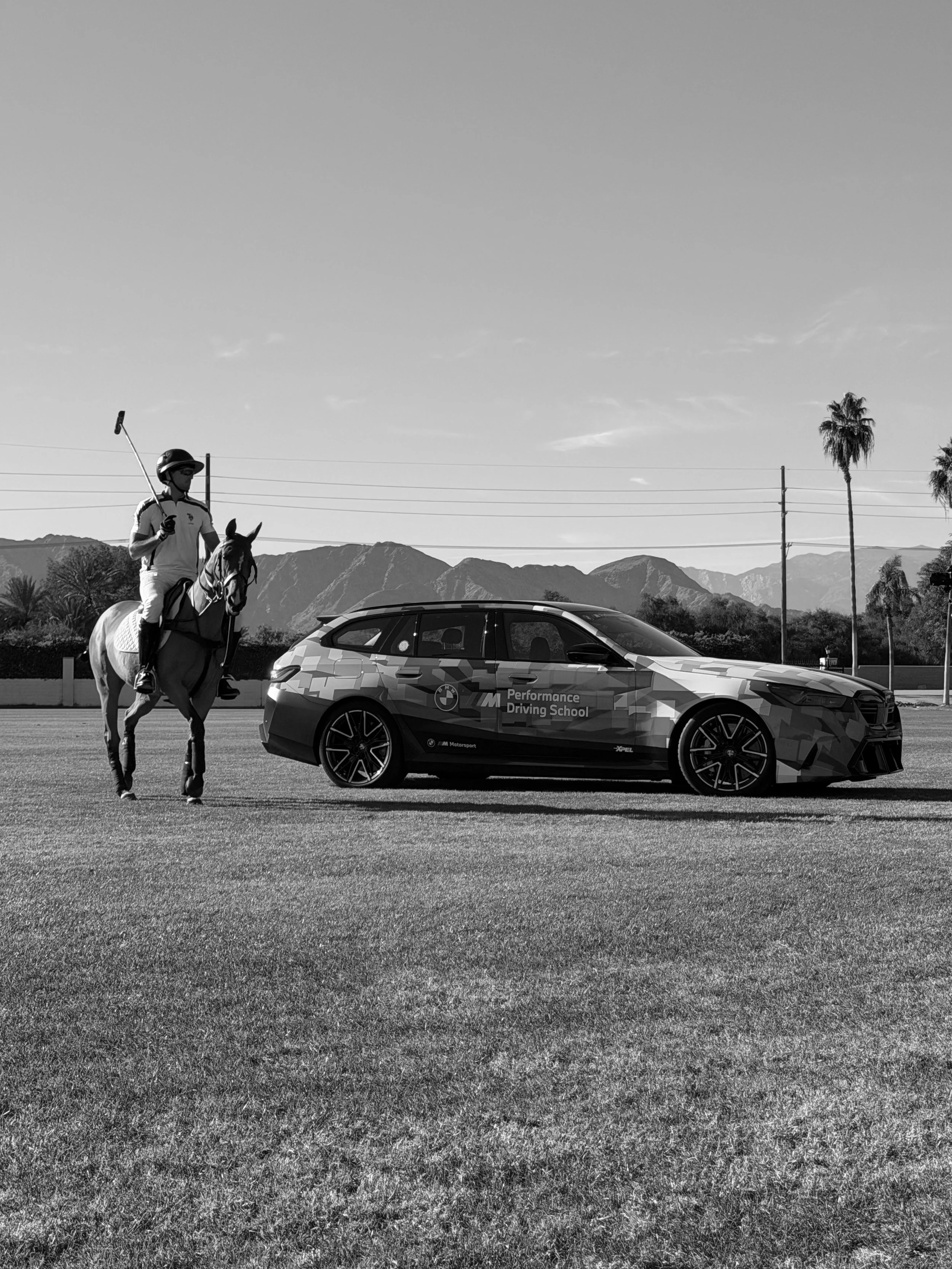 A person on horseback holding a polo mallet stands on a grassy field next to a luxury sports car with "Performance Driving School" written on the side, with mountains and palm trees in the background.