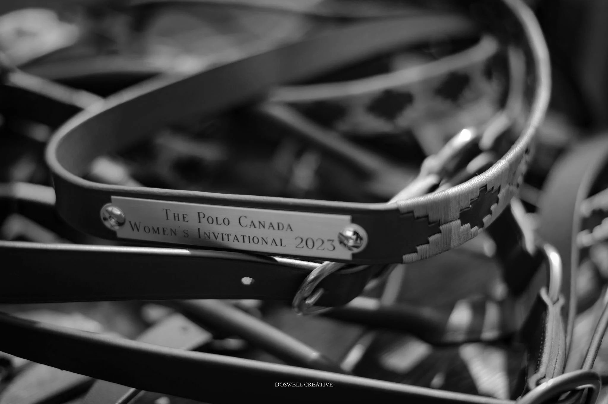 Close-up of a leather halter with an inscription reading "The Polo Canada Women's Invitational 2023" resting on a pile of various belts and accessories.