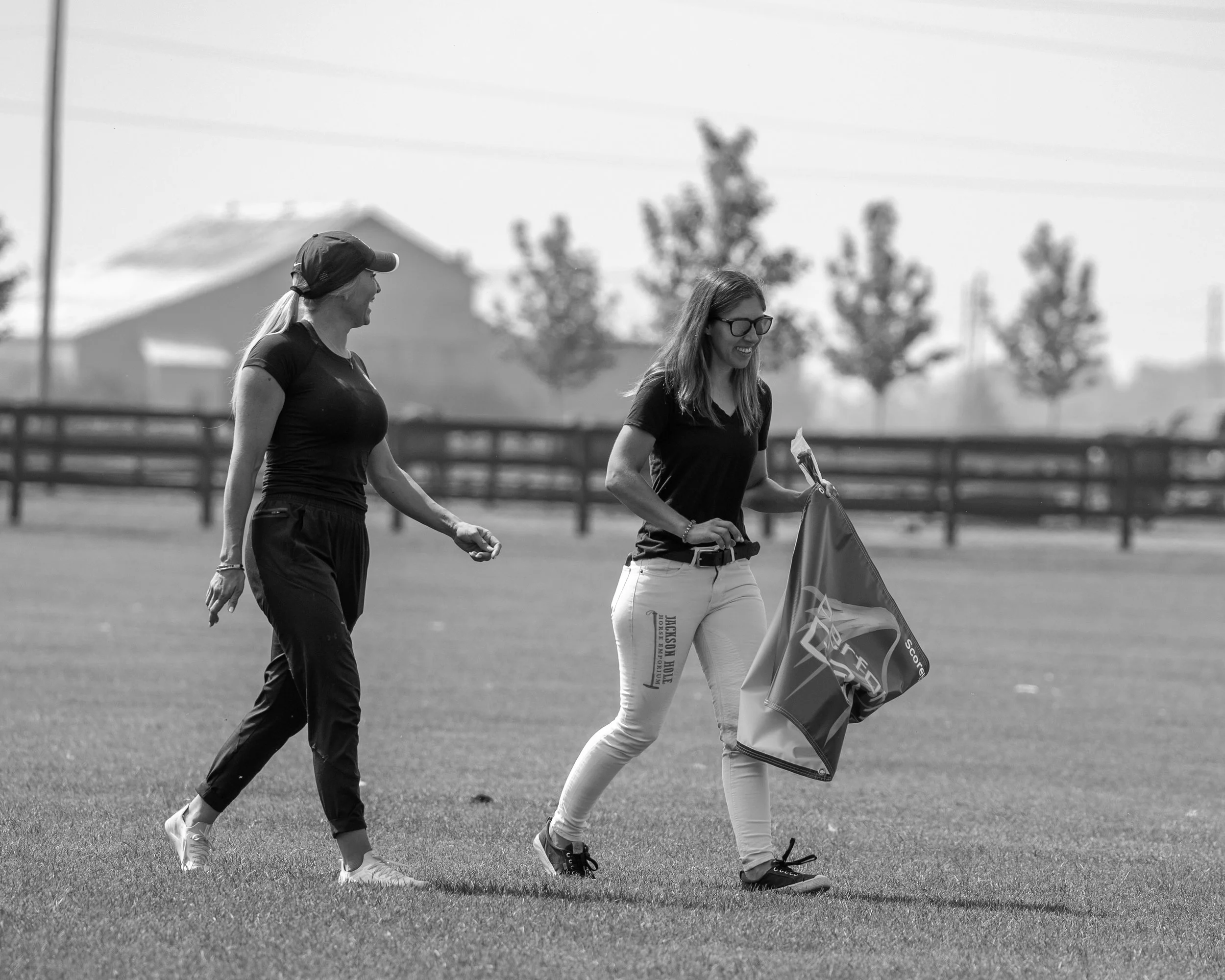 Two women walking across a grassy field, one carrying a folded flag, with trees and a fence in the background.