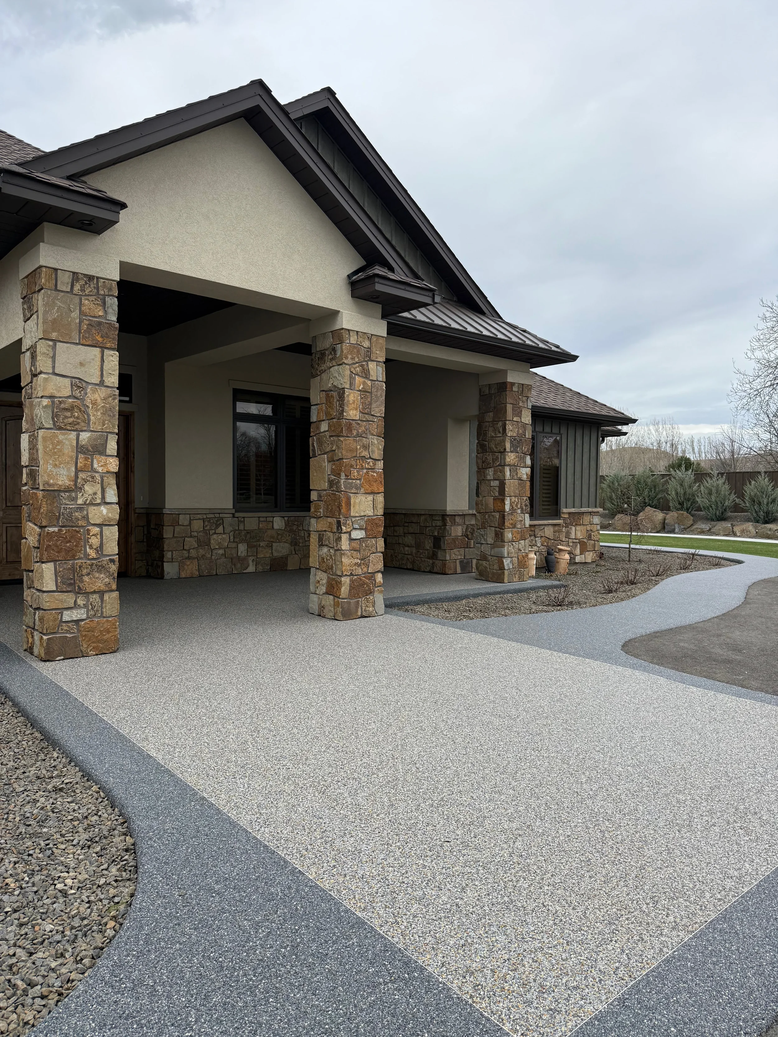 Front view of a house with a stone and stucco exterior, a covered porch, large windows, and a winding concrete pathway in the yard. Resin Bound Surfacing.