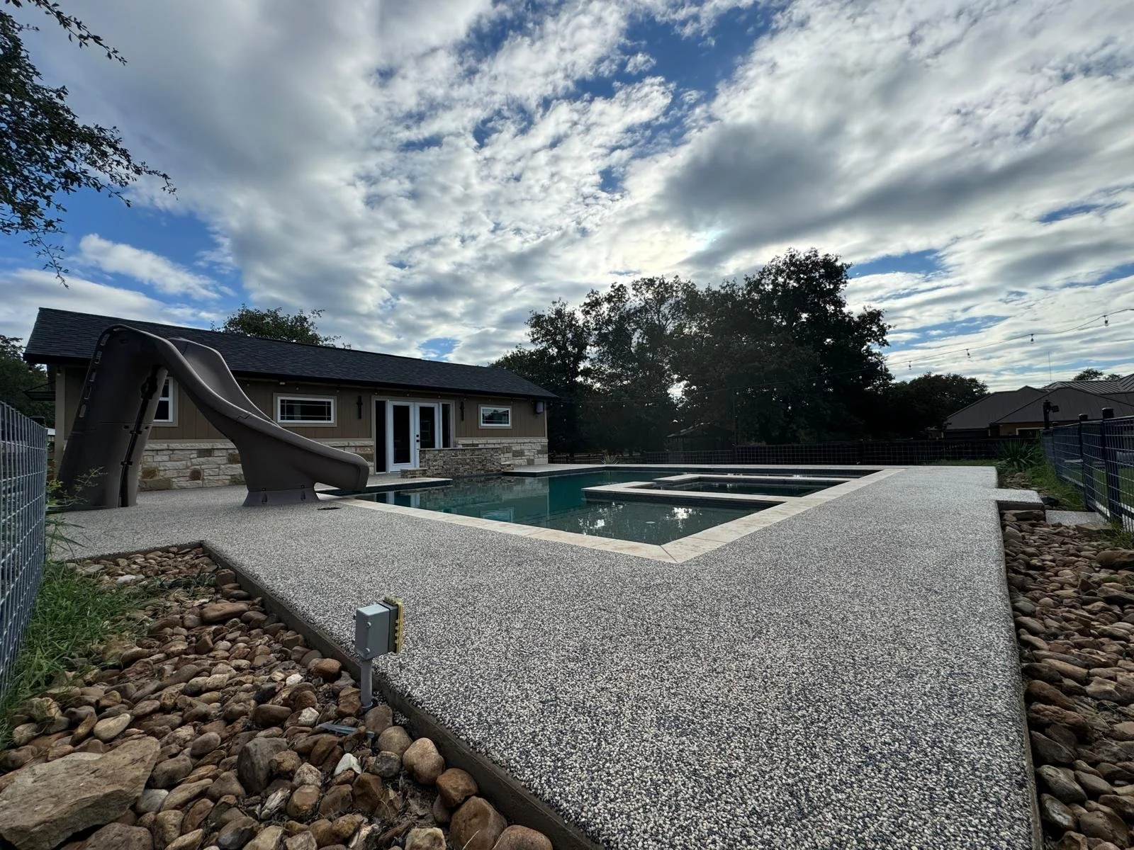 A backyard with a rectangular pool and a water slide, a small building with a stone exterior, and trees under a partly cloudy sky.  Resin Bound Surfacing.