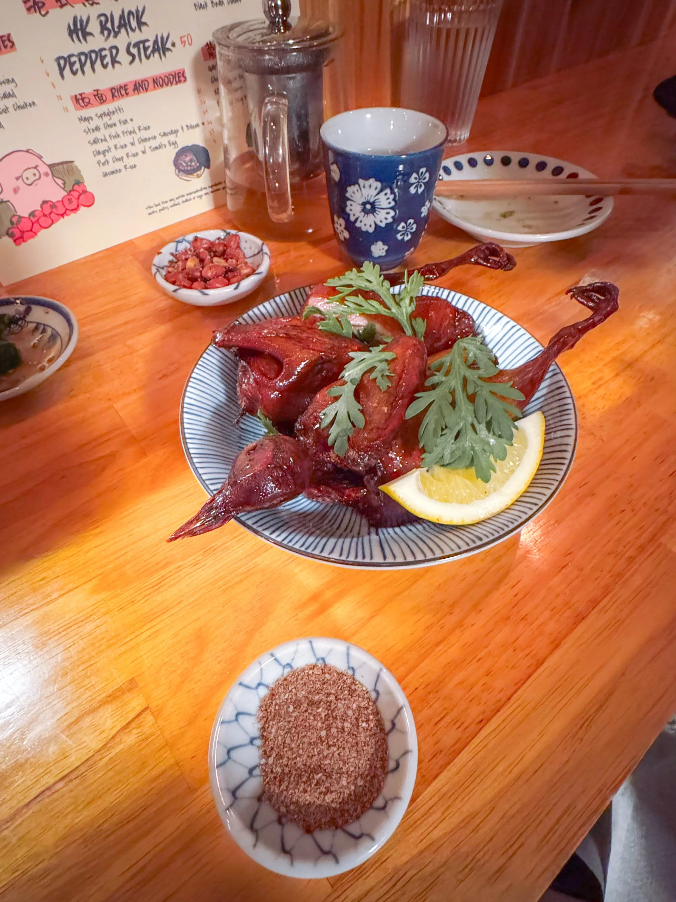 A whole roasted duck garnished with fresh cilantro and lemon wedges, served on a decorative plate on a wooden table. Nearby are small bowls with dipping sauces, and in the background, there is a menu, a glass of water, a blue floral cup, and a clear drinking glass.