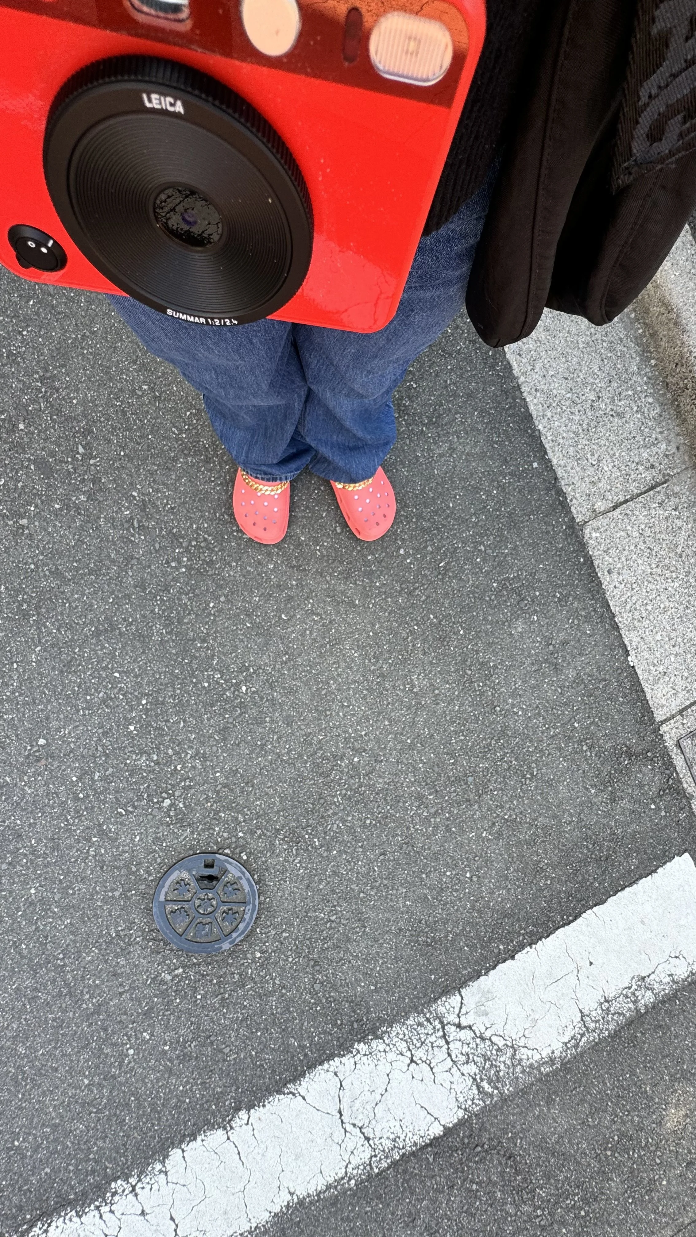 Person standing on asphalt with a white painted parking space line, wearing pink Crocs, blue jeans, black jacket, holding a red portable speaker.